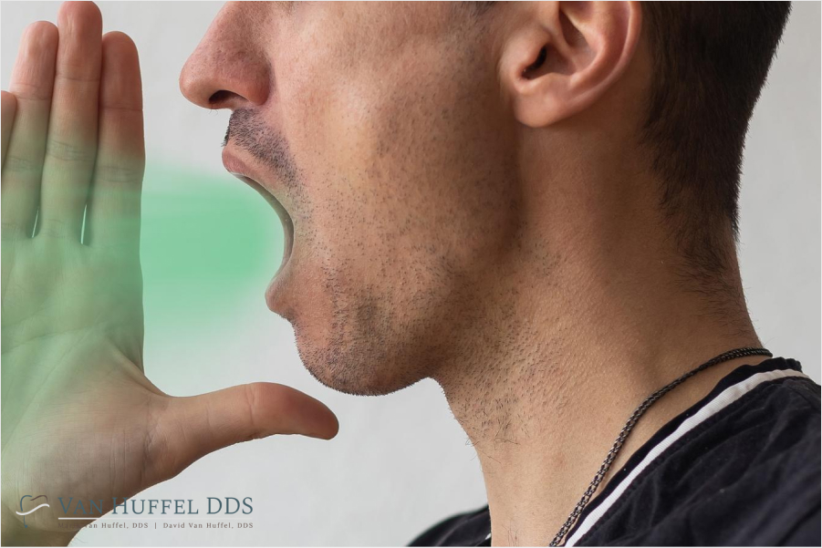 A man with facial stubble cups his hand near his mouth and exhales, with a green mist digitally added to represent bad breath. The image includes a logo for Van Huffel DDS in the lower left corner.